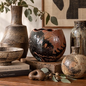 May include: A collection of ceramic vessels including a tall brown vase, a round black and rust-coloured bowl-shaped vase, a textured beige bowl, a dark glazed bottle, and a small round beige vase, with two books titled Australian Studio Pottery.
