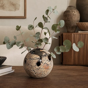May include: A spherical ceramic vase with a textured, cracked beige and brown surface featuring multiple circular openings. The vase contains several stems of green eucalyptus leaves extending upwards and outwards from the top holes.