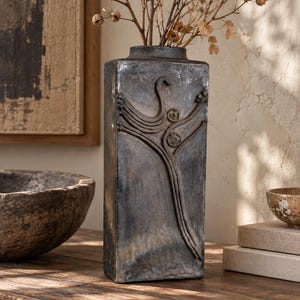 May include: A tall, rectangular, dark grey ceramic vase with a weathered, mottled finish. The front surface features a raised, sculptural relief design of abstract, flowing lines and circular forms. Dried, light brown botanical stems are inside.