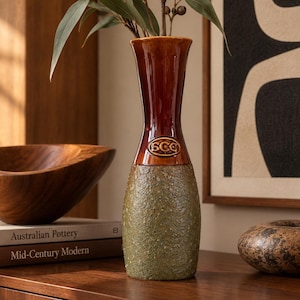 May include: A tall ceramic vase with a smooth, glossy dark brown neck and a textured, matte olive green base. An oval emblem with the letters SCC is embossed on the front. The vase has a flared rim and contains a sprig of eucalyptus leaves.