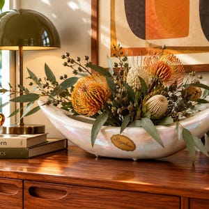 May include: An elongated, iridescent white ceramic bowl with a pointed, boat-like shape. A small, oval-shaped gold plaque with the word "Regalux" is centred on the front. The bowl is filled with a dried floral arrangement of orange protea.