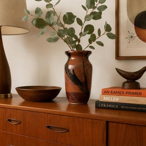 May include: A wooden table lamp with a beige shade, a dark wooden bowl, a ceramic vase with a brown and black glaze containing eucalyptus branches, a dark wooden bird figurine, and three stacked books titled An Eames Primer, Miller, and Ceramics.