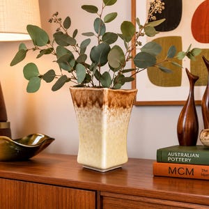 May include: A square ceramic vase with a flared rim, featuring a cream-coloured base that transitions into a dark brown drip glaze at the top. The vase contains a leafy green eucalyptus branch with small round leaves and tiny seed pods.