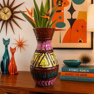 May include: A mid-century modern interior scene featuring a brown, pink, and yellow ceramic vase holding bird of paradise flowers. Other decor includes a lava lamp, cat figurines, a starburst clock, and books.