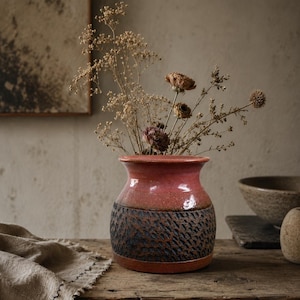 May include: A ceramic vase with a smooth, glossy pink upper section and a textured, dark charcoal-coloured lower section featuring a repeating pattern. The vase contains a collection of dried flowers with thin stems and small, muted-coloured blossoms.