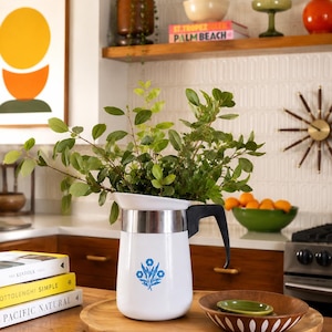 May include: A white coffee pot with a blue floral design, filled with green leafy branches, sits on a wooden tray. The kitchen scene includes a wooden table, books, and a bowl of oranges. An orange pendant light hangs above.