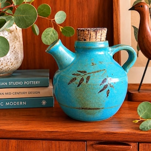 May include: A turquoise ceramic cruet with a rounded body, a side handle, and a short spout. The vessel features a dark brown botanical leaf pattern on the front and is sealed with a cylindrical cork stopper. The glaze has a smooth finish.