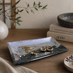 May include: A rectangular ceramic tray featuring a glossy finish with a blend of white, grey, and black abstract streaks. The tray holds a gold chain necklace, a gold ring, and two gold hoop earrings, showcasing a smooth, reflective surface.