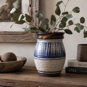 May include: A ceramic vase with a rounded body and a flared rim, featuring a white base with blue vertical brushstroke patterns and speckled details. The vase has a dark brown glazed rim and a blue base, containing green eucalyptus branches.