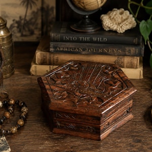 May include: A dark brown hexagonal wooden box featuring intricate hand-carved floral and leaf patterns on the lid and sides. The box has a polished finish and a hinged lid, showcasing detailed craftsmanship with deep grooves and textured wood grain.