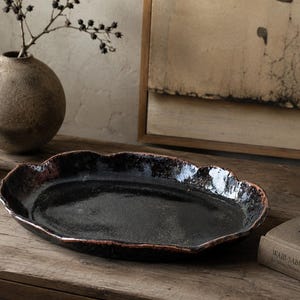 May include: An oval ceramic tray with a glossy black interior and a textured, dark brown rim. The tray features an irregular, organic edge shape with a reflective surface finish, showcasing a deep, dark colour palette and a handcrafted appearance.