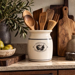May include: A cream-coloured ceramic utensil crock featuring two thin dark blue horizontal bands and a central oval logo with the text Byfield Pottery Queensland Australia, filled with various wooden spoons, spatulas, and a pastry brush.