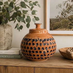 May include: A round, glazed ceramic jar with a lid, featuring a warm orange-brown base colour and a repeating pattern of dark blue, circular indentations across the lower body. The jar has a short, narrow neck and a small, flat-topped lid.