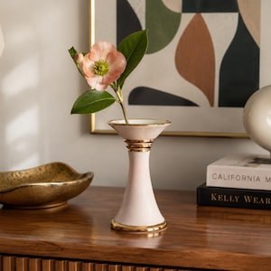 May include: A pale pink ceramic bud vase with a flared top and base, featuring metallic gold trim on the rim, the centre neck, and the bottom edge. A single light pink flower with green leaves is placed inside the narrow opening of the vase.