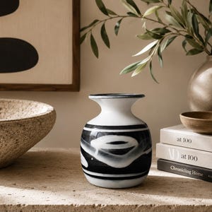 May include: A small, rounded ceramic vase featuring a white base with bold, horizontal black brushstroke patterns. The vase has a narrow neck and a flared rim, with a smooth, glossy finish that contrasts with the hand-painted black markings.