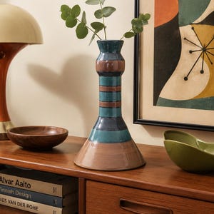 May include: A tall ceramic vase with a flared base and a cylindrical neck, featuring horizontal bands of teal and brown glaze. The vase has a smooth, glossy finish and contains a small green leafy branch extending from the top opening.