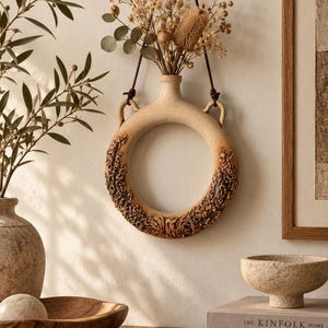 May include: A circular, beige ceramic wall vase with a textured, relief-patterned lower section and two side handles. A dark brown leather cord is attached to the handles for hanging. The vase contains a bouquet of dried flowers and grasses.