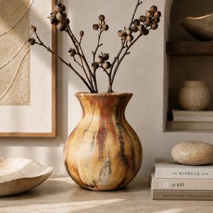 May include: A round ceramic vase with a flared rim, featuring a mottled finish in shades of amber, brown, and cream. The vase contains a cluster of thin, dark brown branches with small, round, dried seed pods attached to the ends of the stems.