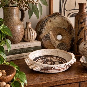 May include: A shallow, round ceramic dish with two side handles, featuring a speckled cream glaze and dark brown hand-painted circular motifs. The dish is displayed alongside various brown stoneware vases, a round donut-shaped vessel, and books.