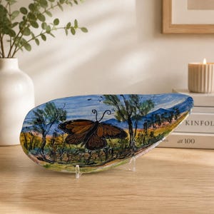 May include: Oval ceramic dish, hand-painted with a butterfly design. The butterfly is brown with black detailing, set against a landscape of trees, fields, and mountains. The dish is supported by a clear plastic stand.