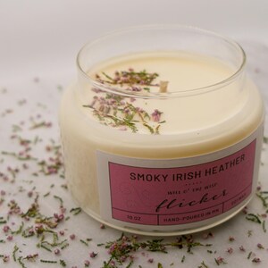 May include: A clear glass jar candle with a creamy wax interior. The candle is labeled "Smoky Irish Heather" and is decorated with small purple flowers. The candle is 10 oz.