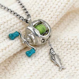 May include: Silver-toned fishing reel pendant necklace with turquoise accents and a fish charm. The reel features green thread and is suspended from a chain. The necklace is displayed on a white, textured surface.