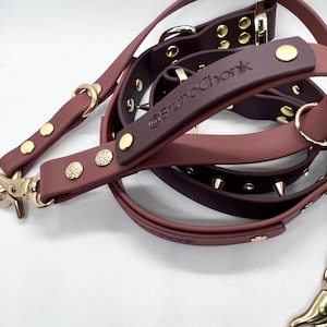 Könnte beinhalten: Eine mauve- und dunkelpflaumenfarbene Hundeleine mit goldfarbenen Metallbeschlägen, darunter Nieten, D-Ringe und Karabinerhaken. Die Leine hat ein eingeprägtes Logo @BrunoChonk am Griff sowie kleine Metallspikes und Nieten.