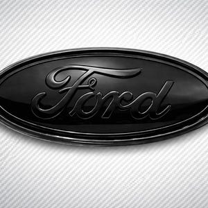 May include: A glossy black Ford emblem with the brand name in a stylized script. The oval-shaped emblem has a shiny, reflective surface and is set against a textured white background.
