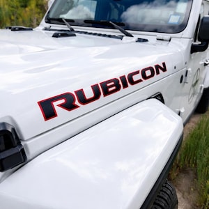 May include: White Jeep Wrangler hood with the word "RUBICON" in black letters with a red outline. The hood is clean and reflects the sky. The front fender and tire are visible.