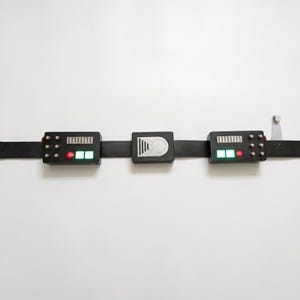 May include: A black utility belt featuring three rectangular modules. The two outer modules have silver vents, small buttons, and illuminated red and green lights. The center module features a silver circular design with vertical lines.