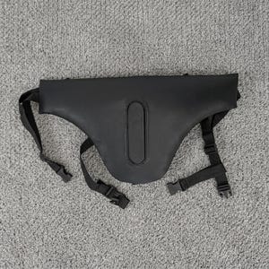 May include: A black, smooth, contoured harness or support accessory featuring a central vertical padded panel. It includes adjustable black nylon webbing straps with plastic side-release buckles extending from the sides and bottom for secure fastening.