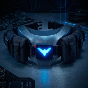 Pode incluir: Um cinto utilitário azul escuro com bolsas e um emblema azul brilhante. O cinto tem uma seção central texturizada e é cercado por várias bolsas pretas. O emblema é uma forma de pássaro estilizada com um detalhe vermelho.