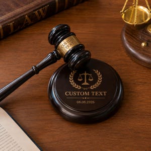 Può includere: Un martello in legno marrone scuro con una fascia in metallo dorato incisa con CUSTOM TEXT, appoggiato su una base circolare in legno abbinata. La base presenta una bilancia della giustizia incisa, CUSTOM TEXT e la data 06.08.2026.
