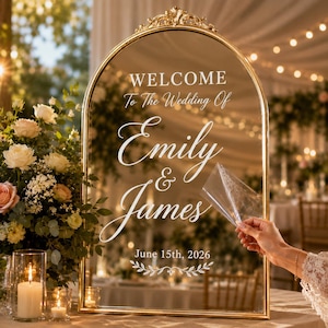 Könnte beinhalten: Ein bogenförmiges Hochzeitsschild aus Spiegelglas mit Goldrahmen und weißem Text Welcome To The Wedding Of Emily & James June 15th, 2026. Eine Hand zieht eine transparente Schutzfolie von der Spiegeloberfläche ab, die einen Raum reflektiert.