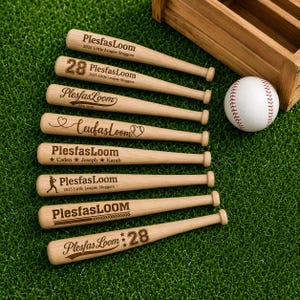 Könnte beinhalten: Acht helle Miniatur-Baseballschläger aus Holz mit verschiedenen individuellen Gravuren, darunter der Text PlesfasLoom, Namen, Zahlen und Sportgrafiken, in einer Reihe neben einem weißen Baseball mit roten Nähten und einer Holzkiste.