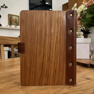 May include: A rectangular notebook with a light brown wood grain cover and a dark brown leather spine. The spine features five evenly spaced brass-colored metal rivets. A small dark brown leather tab is attached to the left side of the cover.