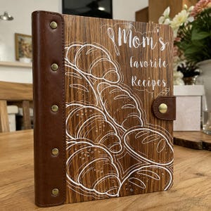May include: A wooden recipe binder with a dark brown leather spine featuring five brass-colored rivets. The front cover displays white line art of various breads and the text "Mom's favorite Recipes" with a whisk icon above the lettering.