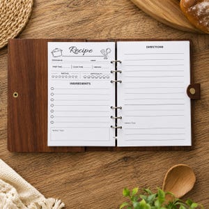 May include: A wooden binder with a dark brown finish and a metal ring mechanism holding white recipe pages. The left page features fields for dish name, date, prep time, cook time, serves, rating, difficulty, ingredients, and notes. The right page is for directions.