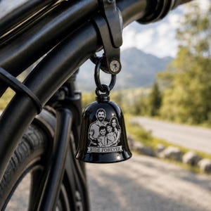 May include: A glossy black bell hanging from a motorcycle frame, featuring a laser-engraved illustration of a family of four. The engraving depicts two adults and two children with the text RIDE FOREVER printed on a banner at the bottom.