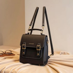 Puede incluir: Una mochila negra estilo cartera con hebillas y herrajes dorados. La bolsa tiene un asa superior y correas ajustables. El texto "KAMIN BAGS" es visible en la parte delantera. Las costuras blancas delinean los bordes de la bolsa.