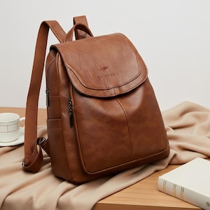 May include: A brown leather backpack with a flap closure and adjustable straps. The bag has a front pocket and side pockets. The brand name is visible on the flap. The backpack is on a wooden surface.
