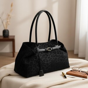 May include: A black handbag with a leopard print pattern. The bag has two long handles and a strap with a gold-colored buckle. The bag is sitting on a cream-colored surface.