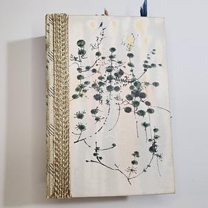May include: A handmade journal with a cream-colored cover featuring a botanical illustration in shades of green and black. The spine is adorned with a gold-colored, woven trim. A blue ribbon serves as a bookmark.