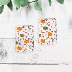 May include: Two clear acrylic charms with a fall-themed pumpkin and floral pattern. The charms are rectangular and have a hole at the top for hanging.