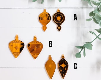 Könnte beinhalten: Eine Sammlung goldfarbener Weihnachtsschmuckstücke in verschiedenen Formen, darunter Tropfen- und Glühbirnendesigns. Jedes Schmuckstück weist Stern- oder Sternmuster auf. Die Ornamente werden vor einem weißen Holzgrund mit grünem Laubwerk präsentiert.