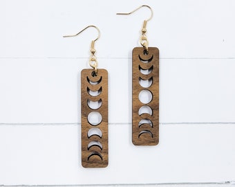 May include: A pair of wooden earrings with a gold hook. The earrings are rectangular and have a design of the phases of the moon cut out.