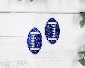 Könnte beinhalten: Zwei blau glitzernde, fußballförmige Ornamente. Jedes Ornament hat einen weißen Streifen oben und unten mit weißen Linien in der Mitte. Die Ornamente befinden sich auf einer weißen Holzoberfläche mit grünen Blättern.