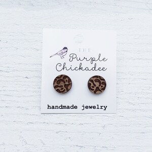 May include: A pair of brown wooden stud earrings with a leopard print design. The earrings are displayed on a white card with the text "The Purple Chickadee handmade jewelry".