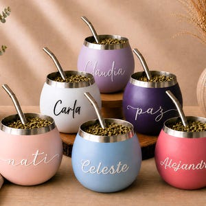 May include: Six round mate gourds with stainless steel rims and metal straws, each filled with dried yerba mate leaves. The gourds are colored light pink, white, lavender, dark purple, light blue, and dark pink, featuring names in script.