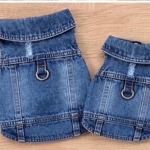 May include: Two blue denim pet vests featuring a fold-down collar, a distressed fabric detail on the upper back, and a silver-toned metal D-ring attachment point centered on the back panel. The vests have visible stitching and a classic denim texture.